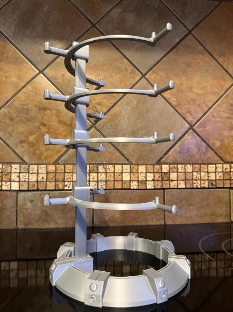 May include: A silver-colored, multi-tiered mug tree with a circular base and multiple arms extending outwards to hold mugs. The tree has a spiral design and is made of a light-colored plastic material. The mug tree is designed to hold multiple mugs.