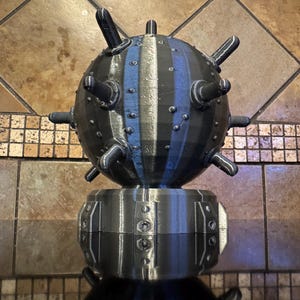 May include: A 3D-printed, black, spherical object with protruding spikes and a striped blue and gray pattern. The sphere sits atop a cylindrical base with riveted details. The object is a decorative piece.