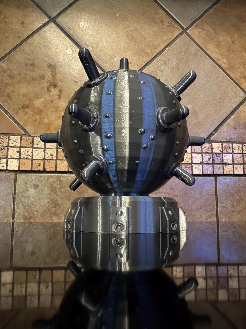 May include: A 3D-printed, black, spherical object with protruding spikes and a striped blue and gray pattern. The sphere sits atop a cylindrical base with riveted details. The object is a decorative piece.
