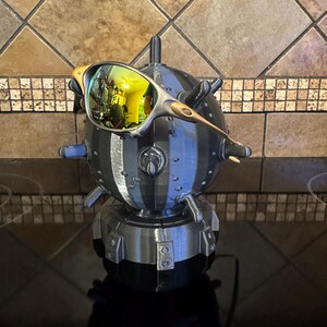 May include: A unique, 3D-printed sculpture of a mine, with a pair of sunglasses resting on top. The sunglasses have a gold frame and reflective lenses. The sculpture is primarily black and gray, with a textured surface and a cylindrical shape.