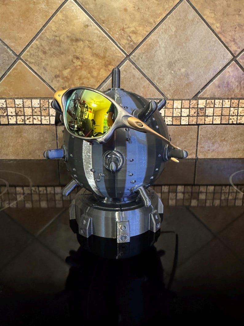 May include: A unique, 3D-printed sculpture of a mine, with a pair of sunglasses resting on top. The sunglasses have a gold frame and reflective lenses. The sculpture is primarily black and gray, with a textured surface and a cylindrical shape.