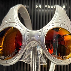 K&ouml;nnte beinhalten: Silberfarbene Sonnenbrille mit orange get&ouml;nten Gl&auml;sern. Die Gl&auml;ser spiegeln die Umgebung wider. Der Text "RESIN OXIDE" ist auf die Gl&auml;ser gedruckt. Die Sonnenbrille wird auf einem transparenten St&auml;nder pr&auml;sentiert.