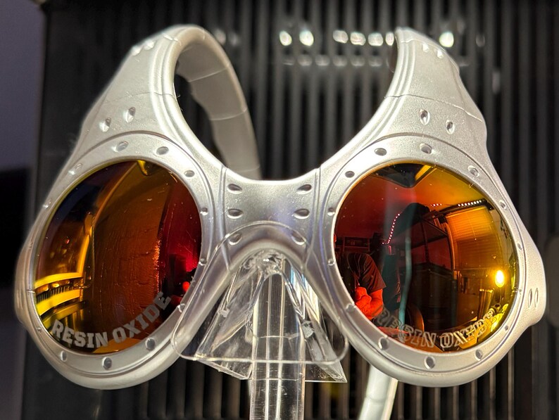 K&ouml;nnte beinhalten: Silberfarbene Sonnenbrille mit orange get&ouml;nten Gl&auml;sern. Die Gl&auml;ser spiegeln die Umgebung wider. Der Text "RESIN OXIDE" ist auf die Gl&auml;ser gedruckt. Die Sonnenbrille wird auf einem transparenten St&auml;nder pr&auml;sentiert.