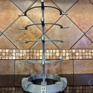 May include: A gray, multi-tiered display stand with a circular base and four curved arms. The stand is made of a matte material and is designed to hold items. The stand is approximately 12 inches tall.