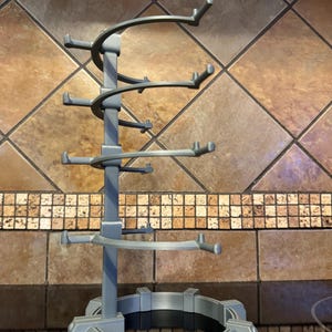 May include: A gray, multi-tiered wine glass holder. The holder has a circular base and a central support structure with curved arms designed to hold multiple glasses. The design is modern and functional.