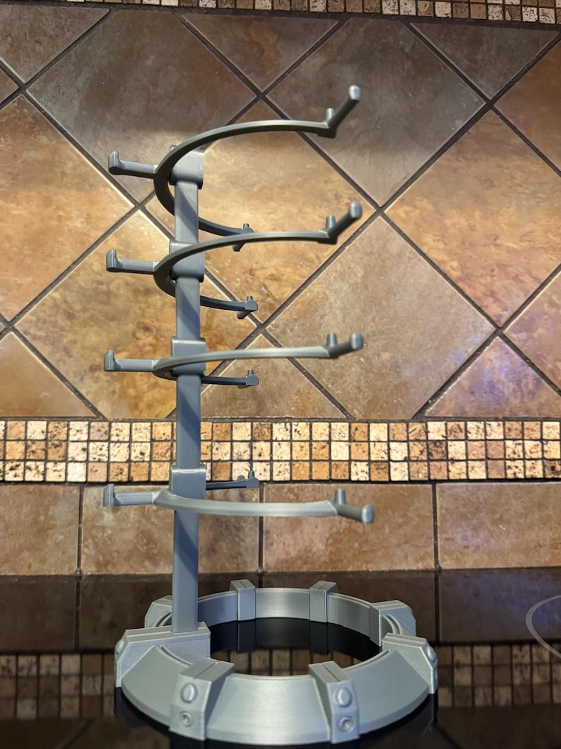 May include: A gray, multi-tiered wine glass holder. The holder has a circular base and a central support structure with curved arms designed to hold multiple glasses. The design is modern and functional.
