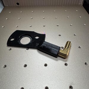 May include: A black, irregularly shaped metal part with a circular opening and two smaller holes. It has a rectangular extension with a brass elbow fitting attached. The part is on a silver surface with evenly spaced holes.