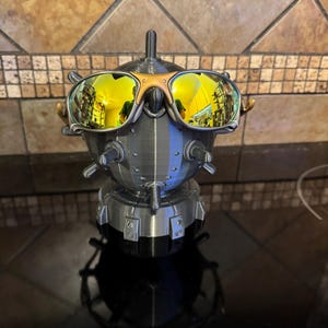 May include: A 3D-printed sculpture of a robot head wearing gold-mirrored sunglasses. The robot is primarily gray and silver, with gold accents on the sunglasses. The sunglasses reflect a yellow hue. The sculpture is on a black surface.