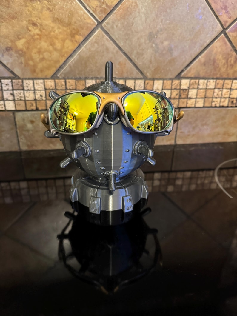 May include: A 3D-printed sculpture of a robot head wearing gold-mirrored sunglasses. The robot is primarily gray and silver, with gold accents on the sunglasses. The sunglasses reflect a yellow hue. The sculpture is on a black surface.
