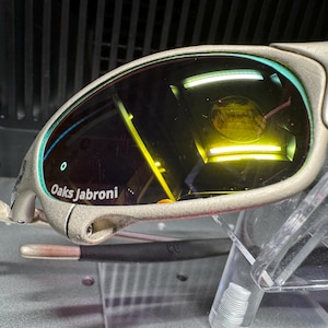 K&ouml;nnte beinhalten: Nahaufnahme einer Sonnenbrille mit silbernem Rahmen und gelb get&ouml;nten Gl&auml;sern. Der Text "Oaks Jabroni" ist auf dem Rahmen zu sehen. Die Sonnenbrille wird auf einem transparenten Kunststoffst&auml;nder pr&auml;sentiert.