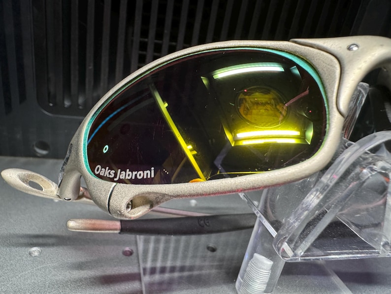 K&ouml;nnte beinhalten: Nahaufnahme einer Sonnenbrille mit silbernem Rahmen und gelb get&ouml;nten Gl&auml;sern. Der Text "Oaks Jabroni" ist auf dem Rahmen zu sehen. Die Sonnenbrille wird auf einem transparenten Kunststoffst&auml;nder pr&auml;sentiert.