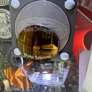 K&ouml;nnte beinhalten: Nahaufnahme eines reflektierenden, kreisf&ouml;rmigen Objekts mit dem Text "Oaks Jabroni" in einer stilisierten Schriftart. Das Objekt ist goldfarben und reflektiert eine Hand, die ein Friedenszeichen macht. Der Hintergrund enth&auml;lt weitere kreisf&ouml;rmige Objekte und das Wort "Oakley".