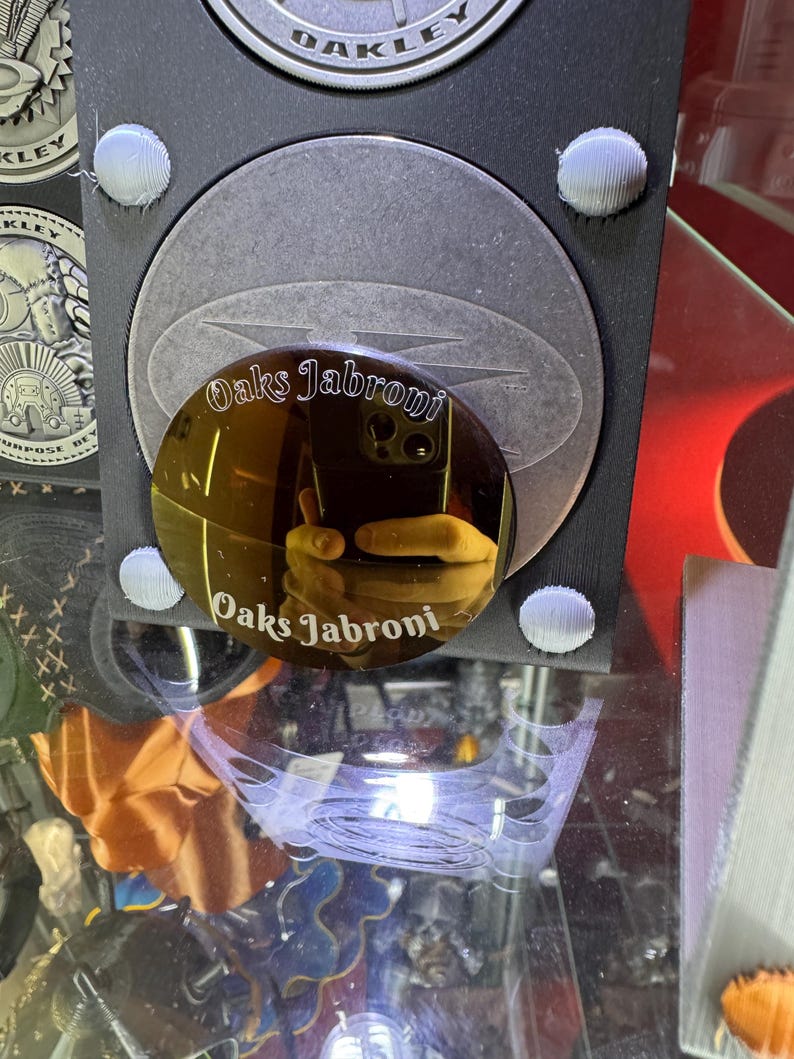K&ouml;nnte beinhalten: Nahaufnahme eines reflektierenden, kreisf&ouml;rmigen Objekts mit dem Text "Oaks Jabroni" in einer stilisierten Schriftart. Das Objekt ist goldfarben und reflektiert eine Hand, die ein Friedenszeichen macht. Der Hintergrund enth&auml;lt weitere kreisf&ouml;rmige Objekte und das Wort "Oakley".