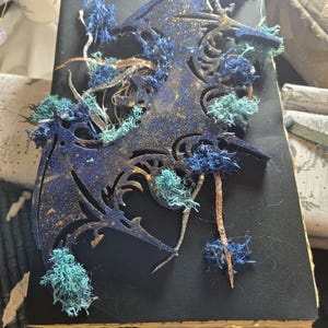 May include: A dark blue journal with a decorative dragon design. The dragon is cut out of the cover and filled with gold glitter. Blue and green moss and glittery branches are attached to the cover.