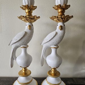 May include: A pair of white and gold candle holders featuring a parrot perched on a pedestal base. The parrot has a textured white body with a gold beak. The candle holder top includes a gold floral accent and a white candle cup.