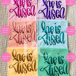 Puede incluir: Seis camisetas de diferentes colores con la frase "He is Risen" en letras doradas, rosas, moradas, verde azulado, oro rosa y verde menta.