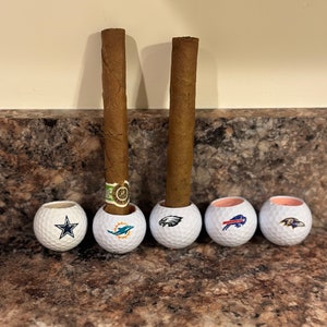 May include: Five golf balls with different NFL team logos are used as cigar rests. Two brown cigars are resting in the golf balls.
