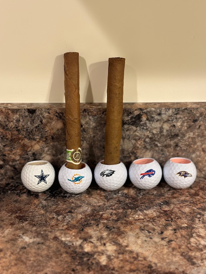May include: Five golf balls with different NFL team logos are used as cigar rests. Two brown cigars are resting in the golf balls.