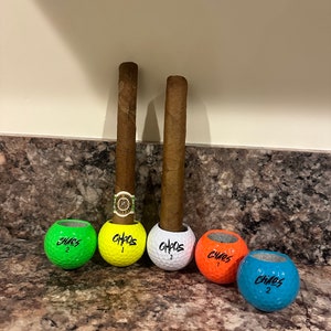May include: Two brown cigars are standing upright in golf ball holders. The golf balls are painted in different colors: green, yellow, white, orange, and blue. Each golf ball has the word "CHAOS" written on it.
