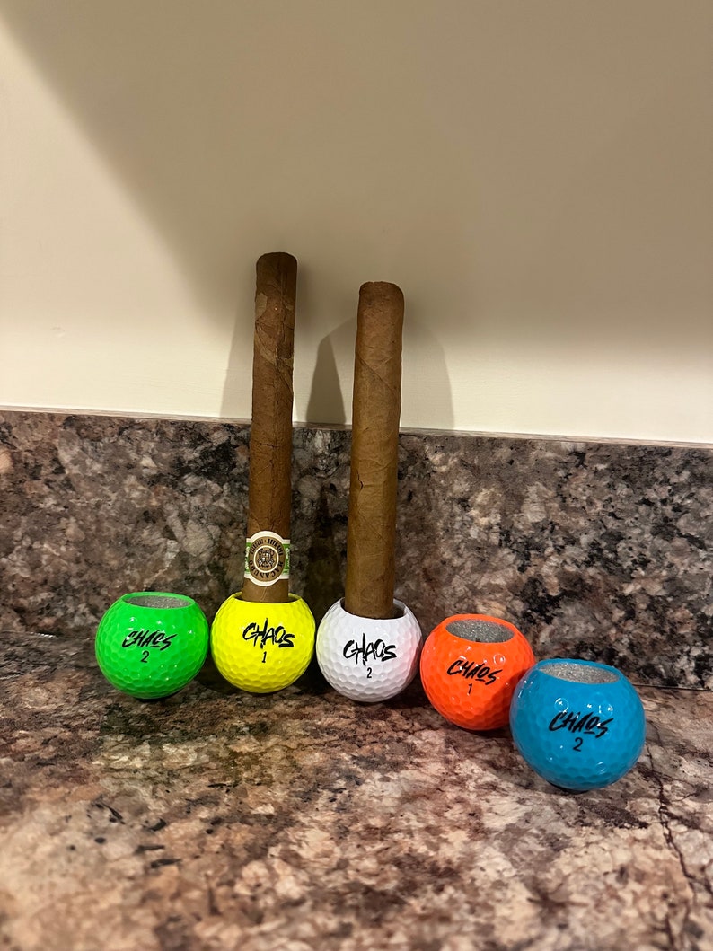 May include: Two brown cigars are standing upright in golf ball holders. The golf balls are painted in different colors: green, yellow, white, orange, and blue. Each golf ball has the word "CHAOS" written on it.
