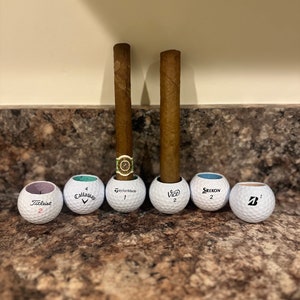 May include: Six golf balls with different brand names, including Titleist, Callaway, TaylorMade, Vice, Srixon, and a ball with a 'B' logo. Two brown cigars are standing upright in the golf balls.