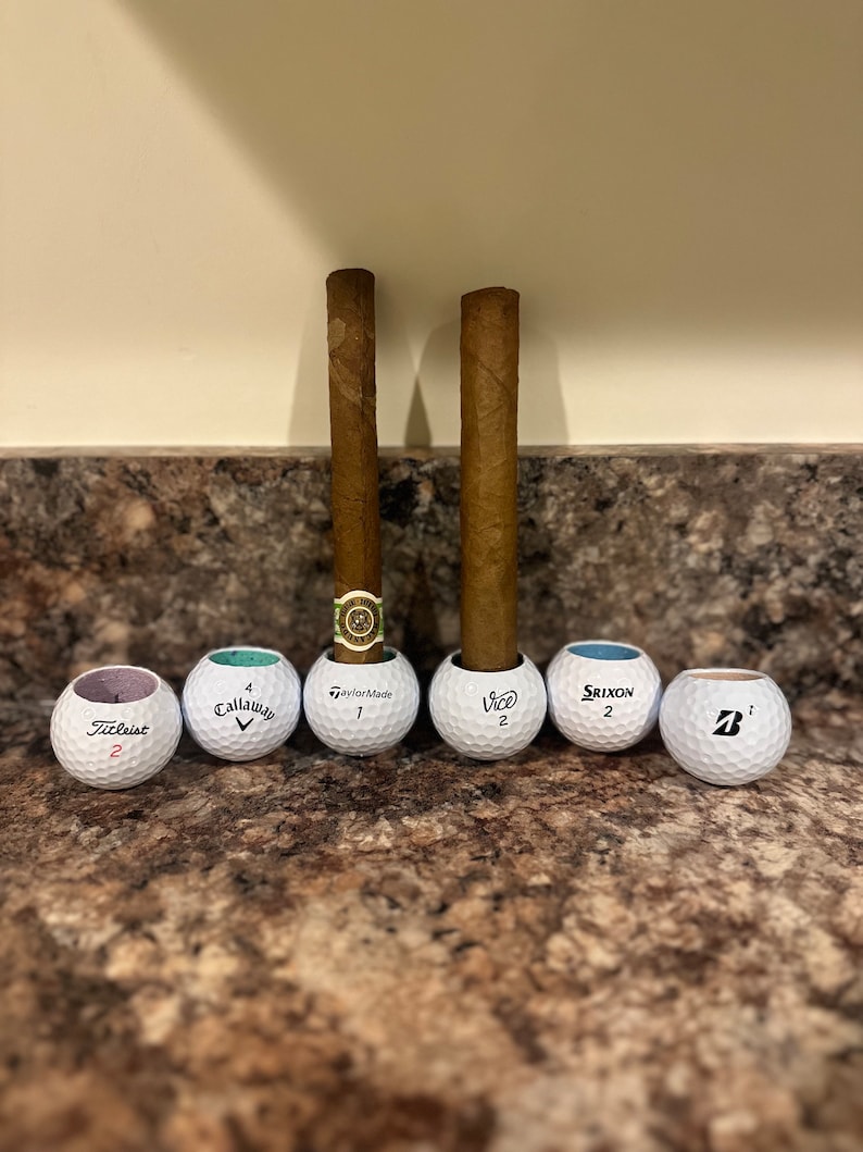 May include: Six golf balls with different brand names, including Titleist, Callaway, TaylorMade, Vice, Srixon, and a ball with a 'B' logo. Two brown cigars are standing upright in the golf balls.