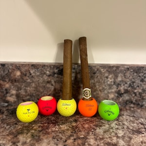 May include: Four golf balls, two yellow, one red, and one green, with the brand names "Top Flight", "Callaway", "Titleist", and "Callaway" printed on them. Two brown cigars are placed in the golf balls.