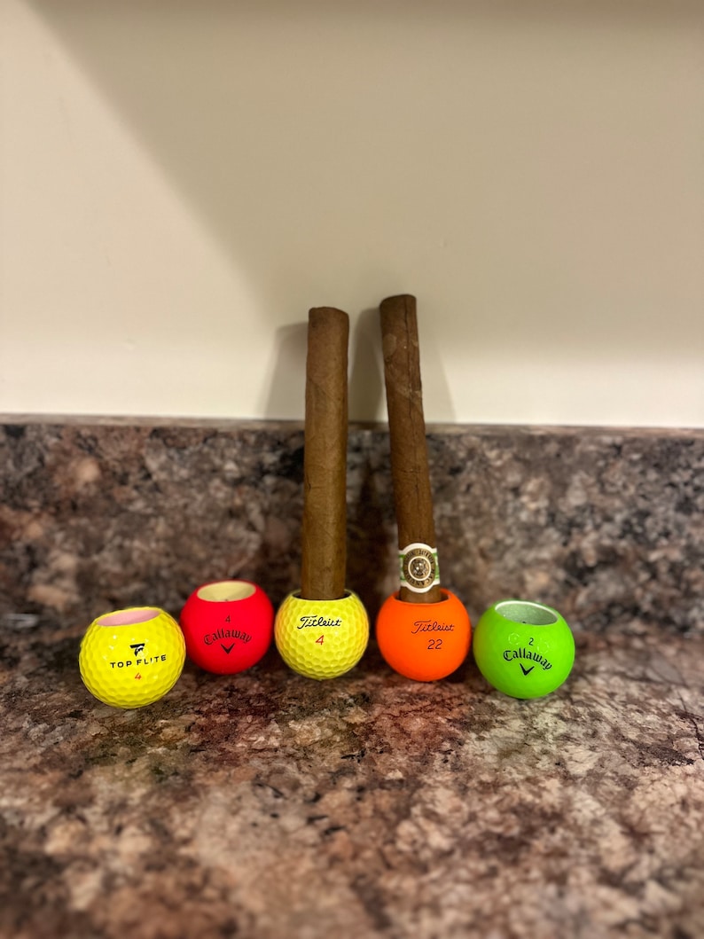 May include: Four golf balls, two yellow, one red, and one green, with the brand names "Top Flight", "Callaway", "Titleist", and "Callaway" printed on them. Two brown cigars are placed in the golf balls.