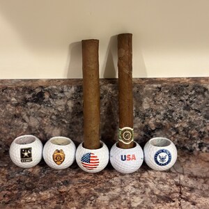 May include: Five white golf balls with different designs. The designs include the US Army logo, a police badge, the American flag, the word "USA", and the US Navy logo. Two brown cigars are standing upright in the golf balls.