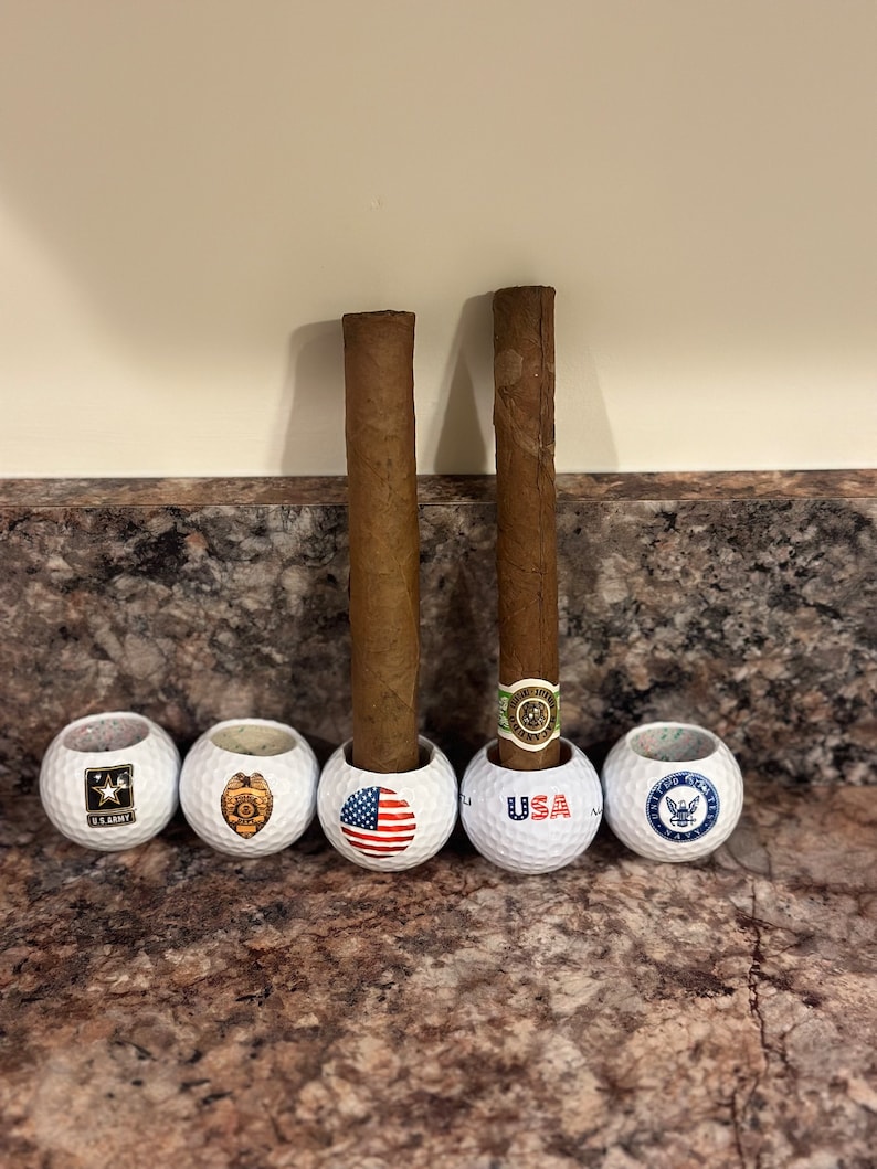 May include: Five white golf balls with different designs. The designs include the US Army logo, a police badge, the American flag, the word "USA", and the US Navy logo. Two brown cigars are standing upright in the golf balls.