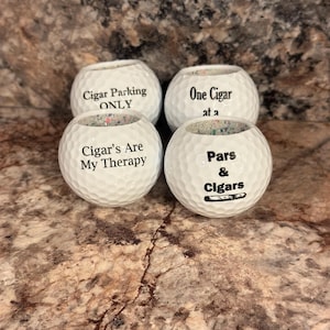 May include: Four white golf balls with black text. The text on the golf balls reads "Cigar Parking ONLY", "One Cigar at a", "Cigar's Are My Therapy", and "Pars & Cigars".
