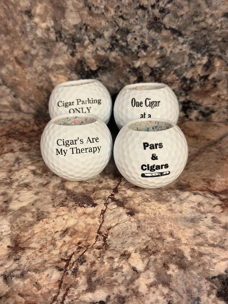 May include: Four white golf balls with black text. The text on the golf balls reads "Cigar Parking ONLY", "One Cigar at a", "Cigar's Are My Therapy", and "Pars & Cigars".