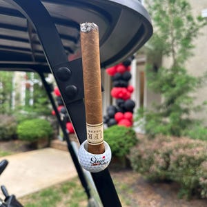 May include: A brown cigar with a tan band labeled JFR is held upright in a white golf ball holder with the text Stogie Golf printed in red and black. The holder is attached to the black metal frame of a golf cart, with a lit ash at the top.