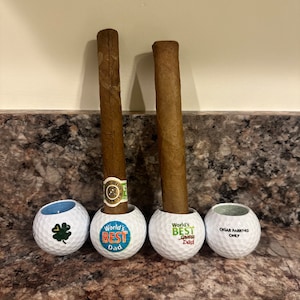 May include: Two brown cigars resting in white golf ball holders. The golf balls are decorated with blue and orange text that reads "World's Best Dad" and "World's Best Golf Dad". The third golf ball has the text "Cigar Parking Only".