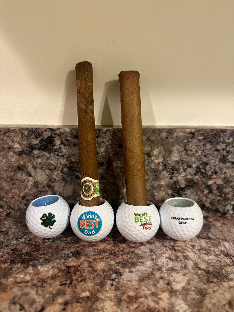May include: Two brown cigars resting in white golf ball holders. The golf balls are decorated with blue and orange text that reads "World's Best Dad" and "World's Best Golf Dad". The third golf ball has the text "Cigar Parking Only".