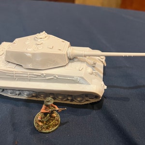 Puede incluir: Maqueta de tanque gris claro con un cañón largo y una figura de soldado en miniatura. El tanque tiene detalles, incluyendo orugas y una torreta. El soldado está en posición de combate.