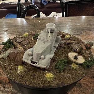 Puede incluir: Maqueta detallada de un coche blindado militar con torreta, ambientada en un diorama con figuras y terreno en miniatura. El modelo es gris, y la base incluye tierra marrón, follaje verde y pequeñas rocas. También hay una figura de soldado.