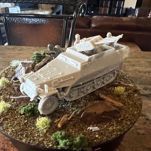 May include: A model of a light grey military half-track vehicle with a mounted gun and miniature soldiers. The vehicle is set on a base with miniature terrain, including rocks, vegetation, and a brown, earthy ground cover.