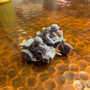 Op de afbeelding: Een grijze miniatuurmotor met zijspan, met twee figuren met helmen. De motor heeft gedetailleerde kenmerken, waaronder een voorwiel, een stuur en een zijspanwiel. De figuren lijken in rijpositie te zitten, wat wijst op een militair thema.