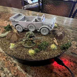 Puede incluir: Un vehículo militar gris en miniatura con seis ruedas y una figura en su interior, colocado sobre una base de diorama con una figura de soldado, follaje y detalles del terreno. La base es redonda y negra, con una luz roja.