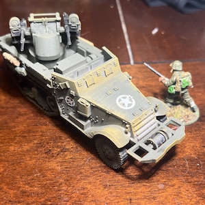 May include: Miniature olive drab military vehicle with a white star emblem and a soldier figure. The vehicle has a rotating turret with two machine guns and a front-mounted winch. The soldier is holding a rifle.