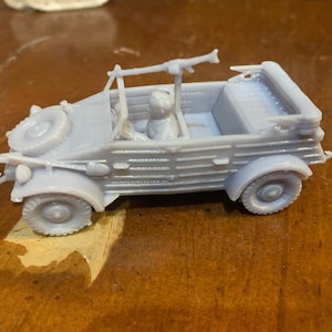 Op de afbeelding: Een lichtgrijze speelgoedauto met een reserveband, koplampen en een machinegeweer bovenop. De auto heeft een bestuurdersfiguur binnenin. Het voertuig is een miniatuurmodel, waarschijnlijk een verzamelobject of een speelgoed.