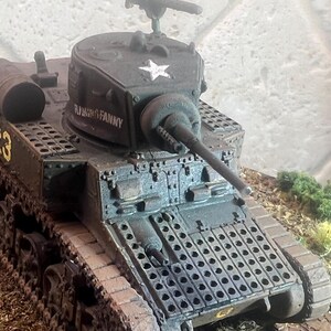 May include: A detailed model tank in olive green with the text "RISING FANNY" and a white star. The tank has a long barrel, a cylindrical side component, and the number "B 23" in yellow. The model is set on a textured surface.