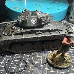 May include: A detailed model of a military tank in olive green with a white star emblem on the turret. A miniature soldier figure with a rifle stands nearby. The scene is set on a textured surface, suggesting a hobby or model-making context.