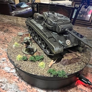 May include: A detailed model of a military tank, painted in olive green, sits on a diorama base with miniature figures and foliage. The tank features a star emblem and a long gun barrel. The base is on a rotating platform.