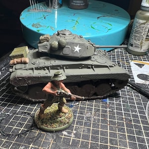 May include: A miniature olive green tank model with a white star emblem, and a miniature soldier figure holding a rifle. The tank has a detailed design with a small storage box. The soldier is standing on a circular base with grass.