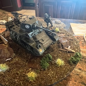 May include: A detailed model of a military tank, painted in olive drab green, with a white star emblem on the turret. The tank is positioned on a diorama base with miniature trees, rocks, and a soldier figurine. The scene evokes a battlefield setting.