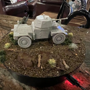 May include: A gray model of an armored car with a rotating base. The model features a turret, gun, and detailed wheels. The base includes miniature terrain with vegetation and small wooden posts. A miniature soldier is visible.