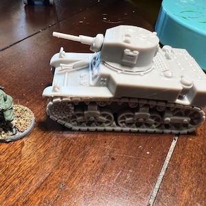 Könnte beinhalten: Ein graues Miniatur-Panzermodell mit langem Lauf und detaillierten Ketten. Eine kleine, bemalte Figur eines Soldaten in grüner Uniform befindet sich in der Nähe auf einer kreisförmigen Basis. Der Panzer und der Soldat sind wahrscheinlich für ein Tabletop-Spiel.