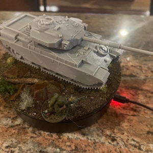 Può includere: Un modello grigio di carro armato con una canna lunga è posto su una base circolare con terreno in miniatura, tra cui erba e una piccola figura. La base è su una superficie marrone, con un cavo di ricarica.